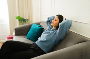 Huzurlu genç bir kadın kulaklıkla müzik dinlemekten hoşlanıyor, rahat bir koltukta gözleri kapalı ve elleri başının arkasında dinleniyor.