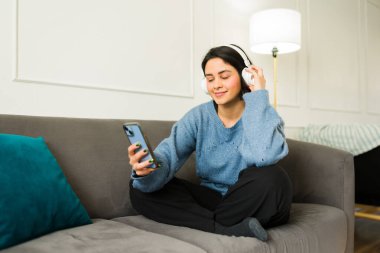 Güzel Latin bir kadın kablosuz kulaklıkla müzik dinlerken evdeki koltukta rahat bir şekilde otururken akıllı telefon uygulaması kullanıyor.