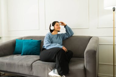Mutlu genç bir kadın rahat koltukta dinleniyor kulaklık takıyor ve evde müzik dinliyor.