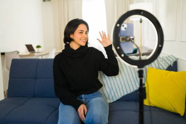 Kadın içerik oluşturucu video blogu kaydediyor, halka ışığının yanında el sallıyor, akıllı telefon ev ortamında vlog içeriğini yakalıyor