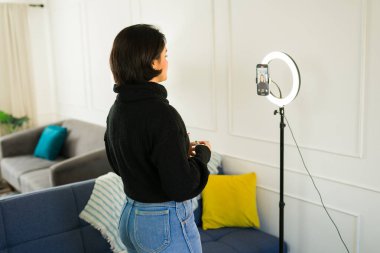 Kadın içerik oluşturucu akıllı telefon kullanarak video blogu kaydediyor, ev ortamında halka ışığının yanında konumlandırılmış, profesyonel görünümlü dijital içerik yaratıyor