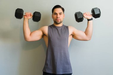 Atletik adam ağır dambıllarla egzersiz yapıyor, gri arka planda omuz egzersizi yapıyor.