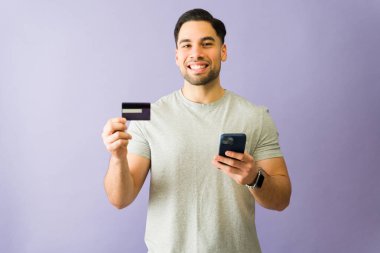 Elinde kredi kartı ve akıllı telefon tutan gülümseyen genç bir İspanyol 'un portresi mor bir arka planda online alışverişe hazır.