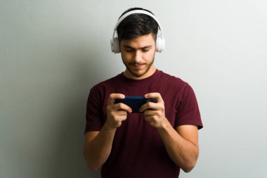 Mobil oyun oynayan, kulaklık takan ve arka planda akıllı telefon tutan genç adam.