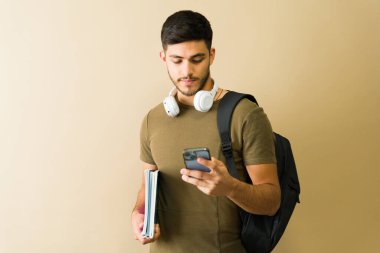 Akıllı telefon kullanan, kitap ve sırt çantası taşıyan odaklanmış bir üniversite öğrencisinin portresi.