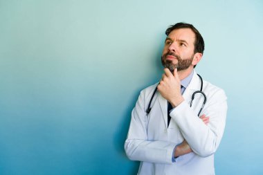 Steteskoplu bir doktor elleri çenesinde düşünür, yukarı bakar ve kollarını eğimli mavi bir arkaplanda kavuşturur.