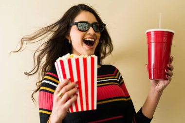 Uzun saçlı neşeli bir genç kadın, 3D gözlük takıyor, elinde patlamış mısır ve içki tutuyor, film izlemekten zevk alıyor.