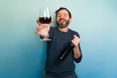 Neşeli sakallı adam elinde şişe tutarken bir bardak kırmızı şarap sunuyor. Mavi arka planda mutlu bir saati kutluyor ve eğleniyor.