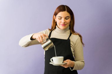 Genç barista kahve bardağına süt dolduruyor, siyah önlük giyiyor, mor arka planda, müşteri için mükemmel bir içecek oluşturuyor.