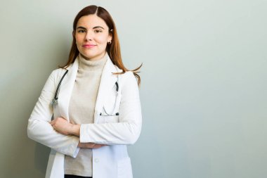 Laboratuvar önlüğü ve steteskop giymiş, açık gri bir arkaplanda kolları çapraz duran kendinden emin bir kadın doktor.