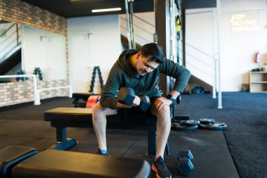 Kilolu orta yaşlı bir adam spor salonundaki bir bankta otururken dambıllarla antrenman yapıyor. Güç antrenmanına ve forma odaklanıyor.