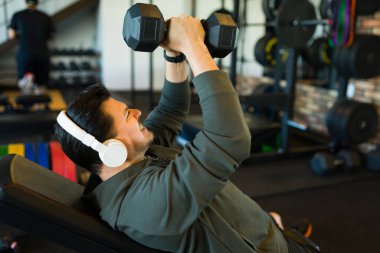 Kas yapan erkek egzersiz yapıyor, kablosuz kulaklık takıyor, modern spor salonunda halter gücü eğitimi veriyor.