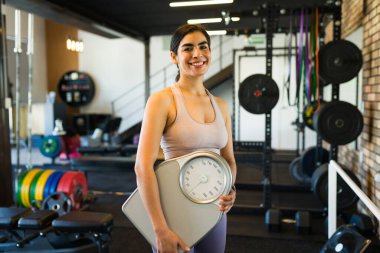 Kilo ölçeği tutarken gülümseyen ve spor salonunda egzersiz yapan neşeli İspanyol kadın spor ve sağlığa olan bağlılığını somutlaştırıyor.