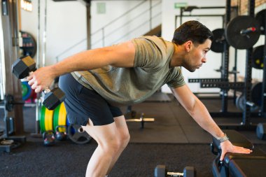 Modern bir spor salonunda yoğun bir egzersiz yaparken ağır dambılları kaldıran kararlı erkek sporcu.