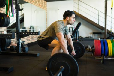 Halter kaldıran bir erkek sporcunun profil görüntüsü, iyi donanımlı bir spor salonunda yoğun bir egzersiz sırasında ağırlık kaldırma hareketleri.
