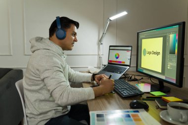 Serbest grafik tasarımcısı logo taslağı yaratıyor, ev çalışma alanında bilgisayar, tablet ve gürültü önleyici kulaklıklarla uzaktan çalışıyor