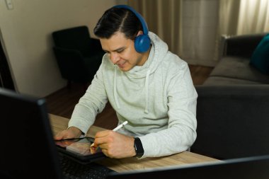 Grafik tasarımcısı kulaklık takıyor ve dijital tablette bir stil kullanıyor, rahat bir oturma odasında evden çalışıyor.