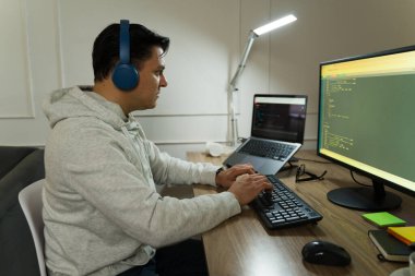 Kulaklık takan yazılım geliştiricisi bilgisayarda kod yazmaya odaklanıyor, merkez ofisten uzaktan çalışıyor.