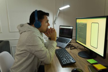 İspanyol yazılım geliştirici daktilo kodu, gürültü önleyici kulaklık takıyor, ev çalışma alanından mobil uygulama oluştururken ekrana odaklanıyor