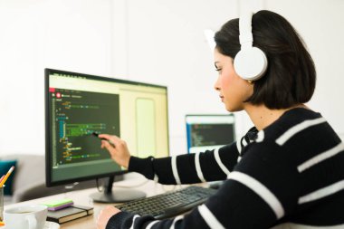 Dizüstü bilgisayarda kulaklık takan kadın programcı, dijital çalışma alanı arka planına sahip programlama arayüzüne işaret ediyor