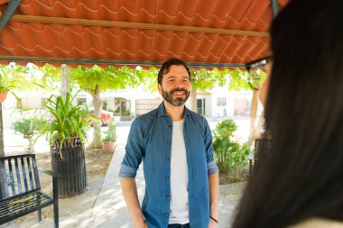 Sakallı neşeli bir adam parkta bir kadınla sohbet ediyor, güneşli bir günün tadını çıkarıyor.