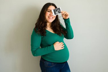 Mutlu hamile kadın, bebeğinin ultrason taramasını gösterirken gülümsüyor ve karnına hafifçe dokunuyor, sevgi ve beklenti yayıyor.