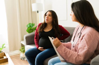 Kadın doktor, hamile bir kadını evinde ziyaret ederken not alıyor, kişiselleştirilmiş sağlık hizmetleri sunuyor ve rahat bir deneyim yaşıyor.