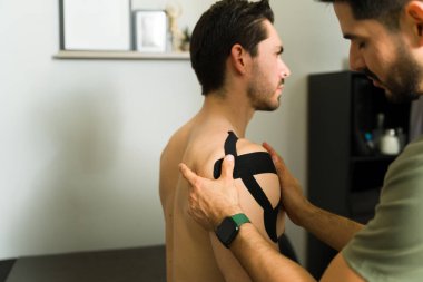 Profesyonel fizyoterapist hastanın omzuna stratejik olarak Kinesio bandı uyguluyor. Ağrı kesicileri ve gelişmiş hareket aralıklarını hedef alıyor.