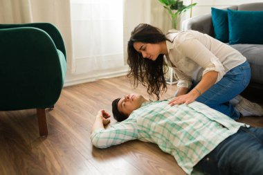 İspanyol kadın, baygın erkek arkadaşının yanında diz çökmüş evde yerde yatıyor, ilk yardım uyguluyor ve hayati belirtileri kontrol ediyor.