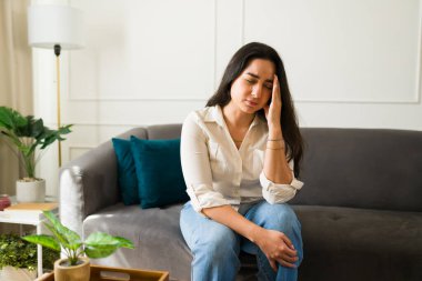 Üzgün latin kadın, evinde, koltuğunda oturup kafasına dokunarak migren ağrısı çekiyor, sağlıksız hissediyor, şiddetli bir baş ağrısı ya da stres yaşıyor.