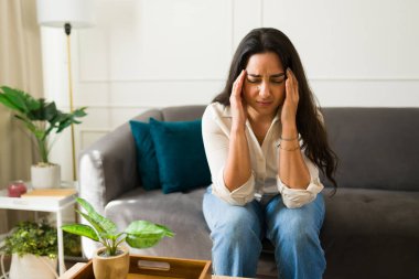Şiddetli baş ağrısı çeken bir kadın. Oturma odasındaki kanepede stresli anlarda migren belirtileri gösteren titreyen şakaklara baskı yapıyor.