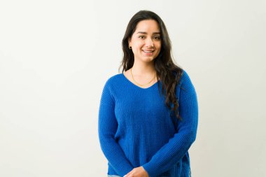Mavi bir süveter giyen, beyaz bir arka plana güvenerek gülümseyen güzel bir Latin kadının portresi.