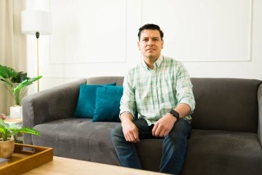 30 'lu yaşlardaki çekici Latin erkek çağdaş kanepede rahatça oturuyor, minimalist yaşam alanında sıcak bir gülümseme yayıyor.