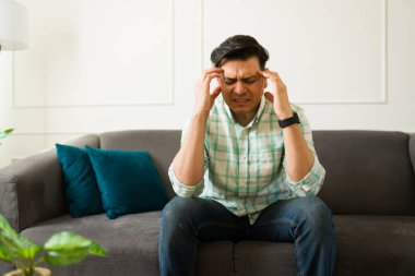 Hispanik erkek şiddetli migren geçiriyor, koltukta otururken tapınak basıyor, yoğun rahatsızlık ve zihinsel gerginlik gösteriyor.