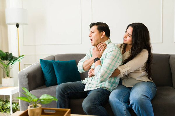 Hispanic woman helping her choking partner by performing the heimlich maneuver in the living room while experiencing a health emergency