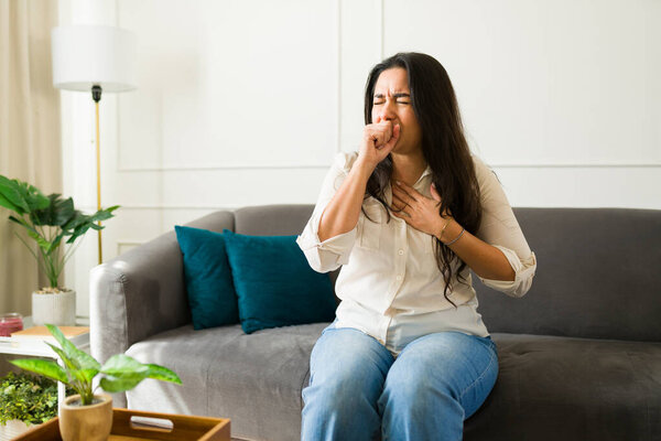 Latin woman wincing from persistent cough, clutching chest while resting on living room sofa, displaying respiratory discomfort