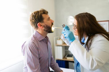 Doktorun muayenehanesinde erkek bir hastanın boğazını muayene eden eldiven takan Latin bir doktor.