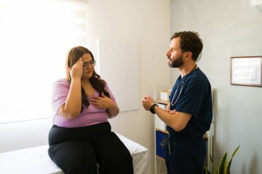 Aşırı kilolu hasta muayene masasında oturuyor ve muayenede doktora belirtileri anlatıyor.