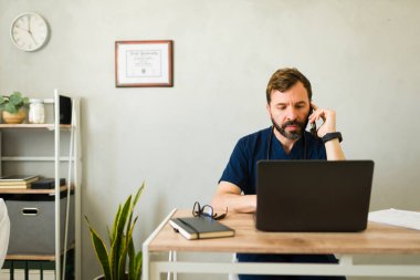 Mavi önlüklü bir tıp uzmanı dizüstü bilgisayarda çalışıyor, klinik ortamda masada otururken telefonla konuşuyor, sağlık hizmetleri desteği dağıtıyor.