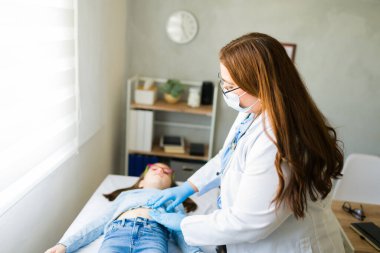 Klinikteki sağlık muayenesi sırasında koruyucu maske takan genç bir hastanın karnını kontrol eden çocuk doktoru.