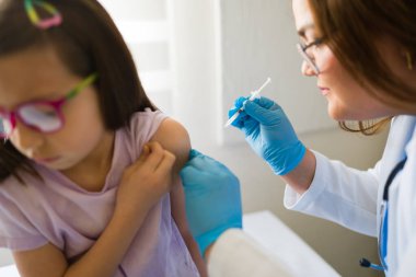 Genç hastaya aşı uygulayan, tıbbi düzende koruyucu giysi giyen kadın çocuk doktoru.