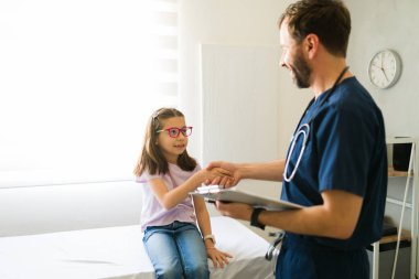 Tıbbi muayeneden sonra gülümseyen erkek çocuk doktoru küçük bir kız hastayla el sıkışıyor.