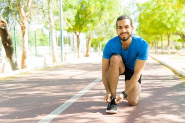 Neşeli bir şehir parkında güneşli bir yaz gününde antrenmana hazırlanmak için koşu ayakkabılarını bağlayan gülümseyen sporcu.