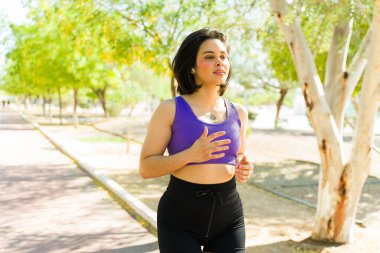 Genç Latin bir kadın parkta canlandırıcı bir koşunun keyfini çıkarıyor, güneşli yaz gününü kucaklarken aktif ve zinde kalıyor.