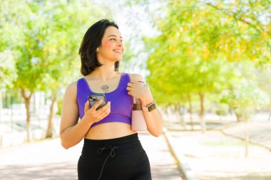 Parkta elinde akıllı telefonu ve su şişesiyle yürüyen gülümseyen sporcu kadın, dışarıda çalışmanın keyfini çıkarıyor.