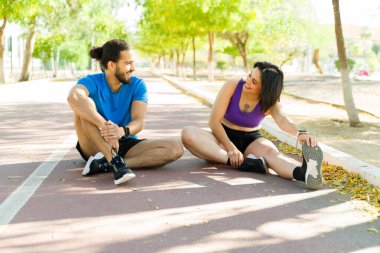 Güneşli bir parkta serinletici bir koşuyu tamamladıktan sonra bacaklarını esnetip gülümseyen mutlu iki sporcu.