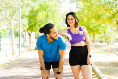 Güneşli bir parkta serinletici bir koşudan sonra, erkek arkadaşını destekleyen gülümseyen İspanyol kadın aktif yaşam tarzlarının tadını çıkarıyor.
