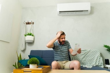 Stresli adam, yaz sıcağında evdeki klima altında koltukta otururken pahalı elektrik faturalarını kontrol ediyor.