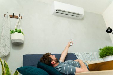 Uzaktan kumandayla kanepede uzanan bir adam yaz sıcağında oturma odasındaki klimayı ayarlıyor.