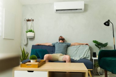 Rahatlamış bir adam, oturma odasındaki rahat bir kanepede uzanırken serin bir klimanın tadını çıkarıyor, yaz sıcağından kaçıyor.